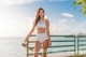 A woman in a white bikini standing on a railing by the water.